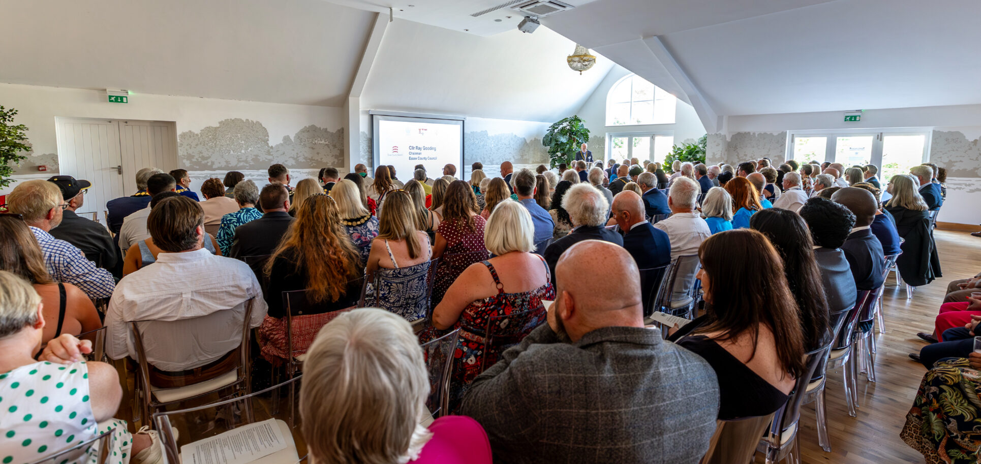 A view from the behind a seated audience in a large room. In the distance a projector screen shows the words Cllr Ray Gooding, Chair, Essex County Council.