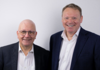 Half portraits of two men smiling at the camera. They both wear jackets and open-necked shirts.