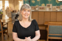 Half portrait of Samantha McReynolds smiling, sitting on a chair in a cafe environment.